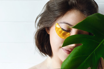 Happy girl with natural skin and lifting anti-wrinkle collagen patches under eyes and green palm leaf. Portrait of beautiful young woman with golden eye patches on white background, copy space