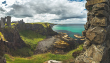 The Overview From Dunluce Castle To The Irish Bay. Overwhelming Northern Ireland Landscape. Epic Shoreline View From Above The Cliff. The Ruins Of The Medieval Building Under The Cloudy Rainy Sky.