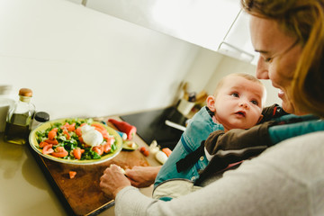 Wall Mural - Smiling baby ported in baby carrier backpack looking at his mother while she cooks, concept of family conciliation