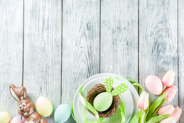 Easter holiday table setting with rabbits and eggs, plates with colorful eggs and chocolate bunny rabbits, festive ribbon, wooden background top view copy space