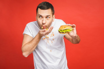 Funny hungry bearded man eating junk food. Excited young man greedily eating hamburgers isolated on red background.