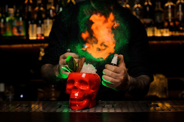 Bartender spraying on the cocktail in the scull cup from the vaporizer in the green light and fire it