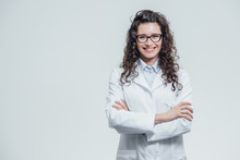 Portrait Of Smiling Young Female Doctor. Beautiful Brunette In White Medical Gown In Glasses. Holding Without A Stethoscope On A Gray Background.