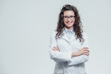 Portrait of smiling young female doctor. Beautiful brunette in white medical gown in glasses. Holding without a stethoscope on a gray background.