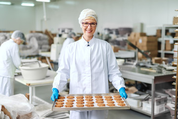 Smiling blonde Caucasian employee in sterile uniform and with eyeglasses standing and holding tray with cookies. Food factory interior. In background other employees working.