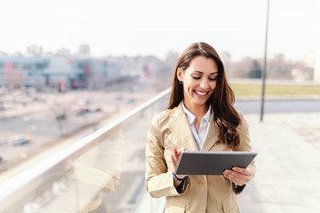 Beautiful caucasian brunette with toothy smile and dressed smart casual using tablet on rooftop.