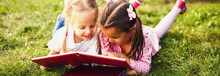 Two Young Girls Lying On The Grass Outdoors Reading Separate Books. These Siblings Are Enjoying Time Together As Family During Summer