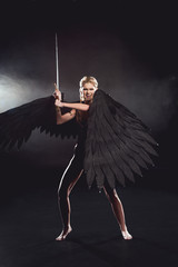 Wall Mural - beautiful angry woman in warrior costume with angel wings holding sword and posing on black background