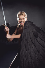 beautiful angry woman in warrior costume with angel wings holding sword and posing on black background