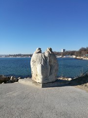 Wall Mural - Large stone rough statue in the form of two fishermen in hats against the blue sea and sky