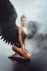 Canvas Print - beautiful sexy woman with black angel wings sitting, looking at camera and posing on dark background