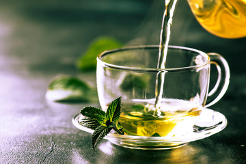 Hot chinese green tea with mint, with splash pouring from the kettle into the cup, steam rises, dark background, selective focus