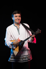 A brunette man in a folk shirt plays a balalaika in scenic blue and red light on a black stage