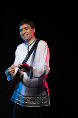 A brunette man in a folk shirt plays a balalaika in scenic blue and red light on a black stage