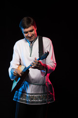 A brunette man in a folk shirt plays a balalaika in scenic blue and red light on a black stage