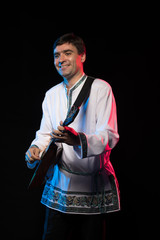 A brunette man in a folk shirt plays a balalaika in scenic blue and red light on a black stage