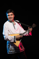 A brunette man in a folk shirt plays a balalaika in scenic blue and red light on a black stage