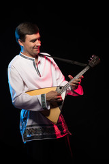 A brunette man in a folk shirt plays a balalaika in scenic blue and red light on a black stage