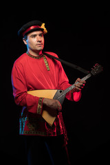 A brunette man in a folk shirt plays a balalaika in scenic blue and red light on a black stage