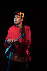 A brunette man in a folk shirt plays a balalaika in scenic blue and red light on a black stage