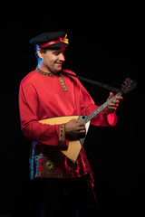 A brunette man in a folk shirt plays a balalaika in scenic blue and red light on a black stage