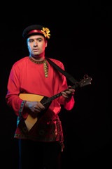 A brunette man in a folk shirt plays a balalaika in scenic blue and red light on a black stage