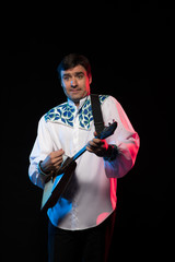 Wall Mural - A brunette man in a folk shirt plays a balalaika in scenic blue and red light on a black stage