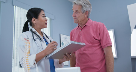 Low-angle view of Japanese woman doctor talking to senior Caucasian male patient. Physician talks to aging man about back problems in her medical practice office