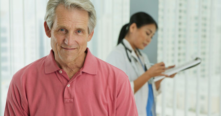 Wall Mural - Close up portrait of senior Caucasian male patient smiling at camera while doctor works in background. Attractive older man in hospital with female physician out of focus behind him