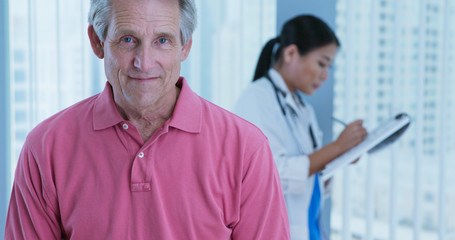 Portrait of Attractive senior man in hospital with female physician out of focus behind him. Older Caucasian male patient looking at camera and smiling while doctor works in background