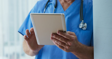 Wall Mural - Close up on Senior Caucasian male doctor to tablet computer as he reads medical history of patient. Older man medical professional looking over records on pad