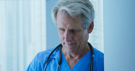 Wall Mural - Close up of senior Caucasian male doctor reading patient notes in hospital. Medical professional looking down. Nurse of surgeon in close-up leaning against pillar