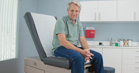 Wall Mural - Portrait of Senior Caucasian male patient sitting on exam room table looking at camera. Older man in hospital for regular check up