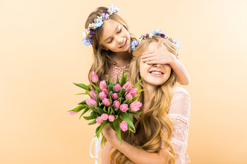 happy mother holding bouquet of pink tulips while cheerful child closing her eyes with hand on yellow background