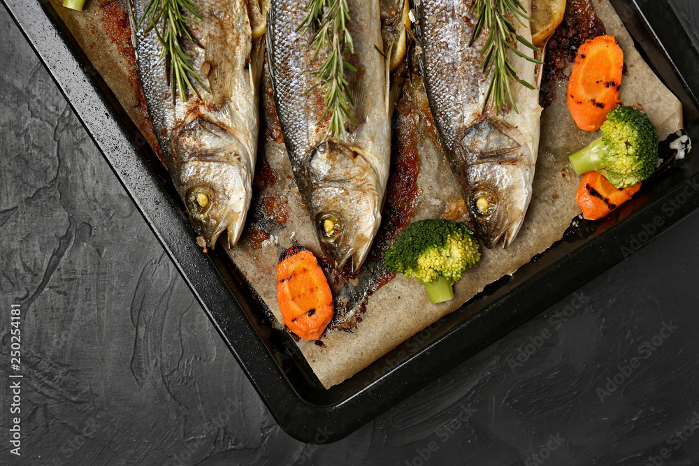 Baking sheet with tasty cooked seabass fish and vegetables on dark table