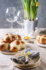 Easter table setting with plate of hot cross buns, narcissus flowers, wine glass on table with linen tablecloth.