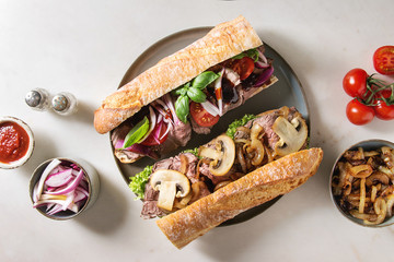 Wall Mural - Variety of beef baguette sandwiches with champignon mushrooms, green salad, fried onion, tomatoes served on ceramic plate with ingredients above over white marble background. Flat lay, space