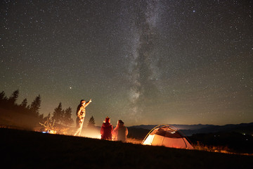 Happy friends hikers having a rest together around campfire beside camp and tourist tent at night in the mountains. Man pointing at incredibly beautiful night starry sky full of stars and Milky way.