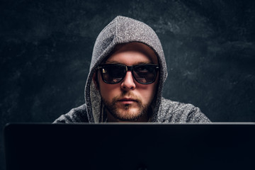 Close-up portrait of a hacker in hoodie and sunglasses sits behind laptop in office against a dark wall 