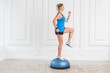 © khosrork - Side view portrait of concentration sporty beautiful young athletic blonde woman in black shorts and blue top working in gym doing exersice in bosu balance trainer, raised one knee on fitness ball.