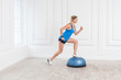 © khosrork - Side view portrait of focused sporty young athletic blonde woman in black shorts and blue top working in gym doing exersice in bosu balance trainer, making one step on fitness ball. Indoor,studio shot