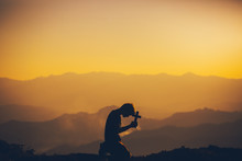 Young Man Kneeling Down Praying And Holding Christian Cross For Worshipping God At Sunset Background. Christian Silhouette Concept.