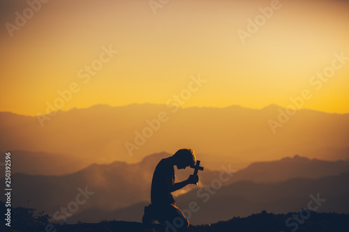 Young man kneeling down praying and holding christian cross for worshipping God at sunset background. christian silhouette concept.