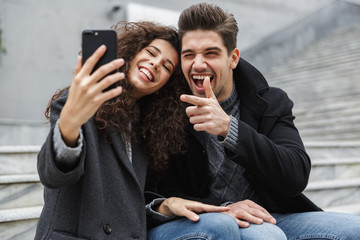 Wall Mural - Image of beautiful couple man and woman 20s in warm clothes, taking selfie photo on cell phone while sitting on stairs outdoor