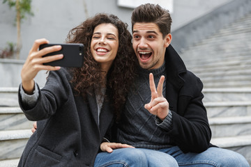 Poster - Image of brunette couple man and woman 20s in warm clothes, taking selfie photo on cell phone while sitting on stairs outdoor