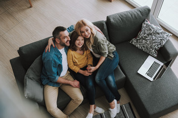 Wall Mural - Happy parents hugging their daughter while sitting on couch with laptop