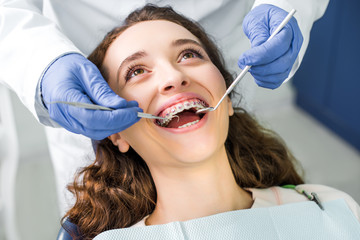 Wall Mural - cropped view of dentist in latex gloves examining cheerful woman in braces with opened mouth