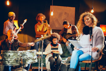 Wall Mural - Jazz band preparing for the gig. In foreground woman singing while the rest of the band playing bass guitar, clavier and acoustic guitar. Home music studio interior.