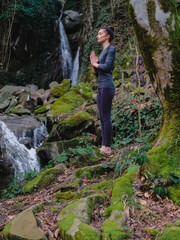 Young slim woman practicing yoga outdoors in moss forest on background of waterfall. Unity with nature concept. Girl standing with prayer hands