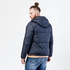 Wall Mural - Fashionable man with beard and bun hairstyle dressed in yellow sweater, jeans and dark down jacket poses in the studio on the white background